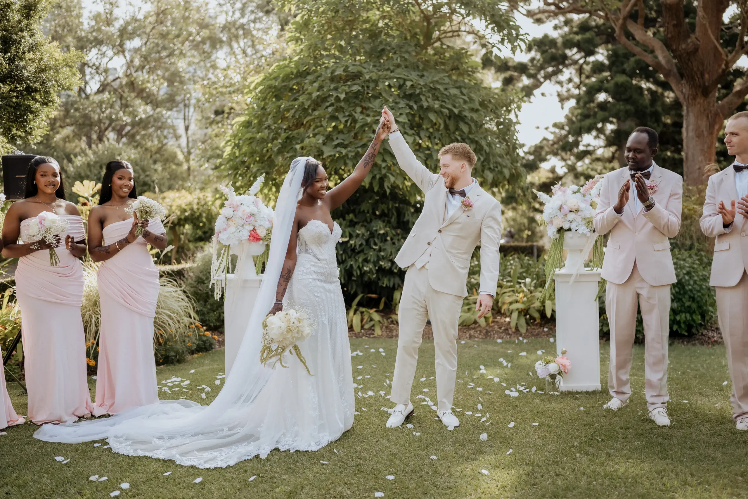 A couple raising their joined hands after a garden ceremony, the groom in cream and the party in blush either side of them, white petals scattered across the grass.